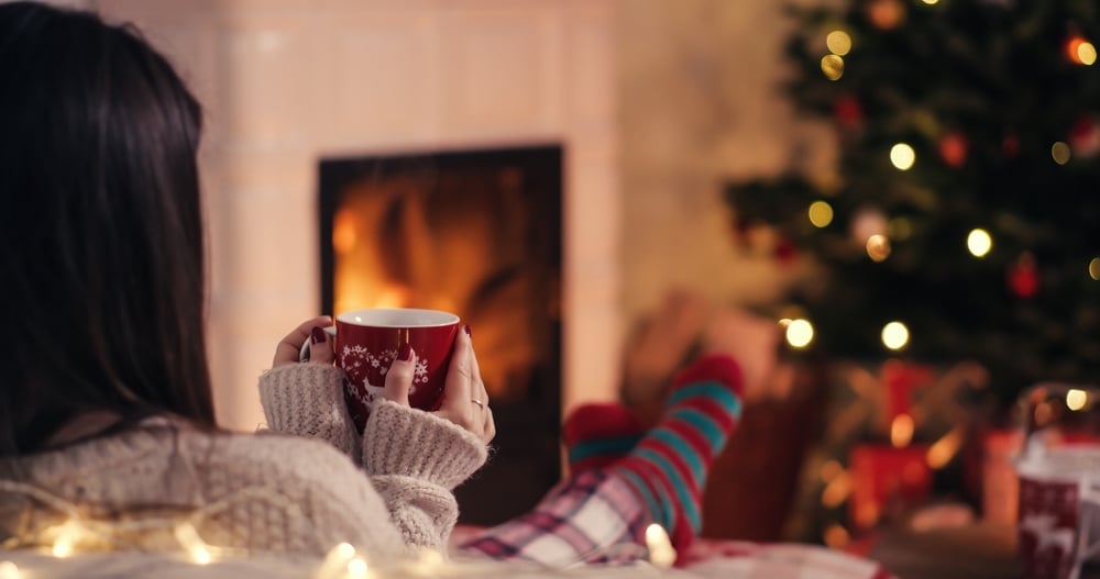 girl with hot drink in a mug overlooking a fireplace and christmas tree practicing self-care during the holidays