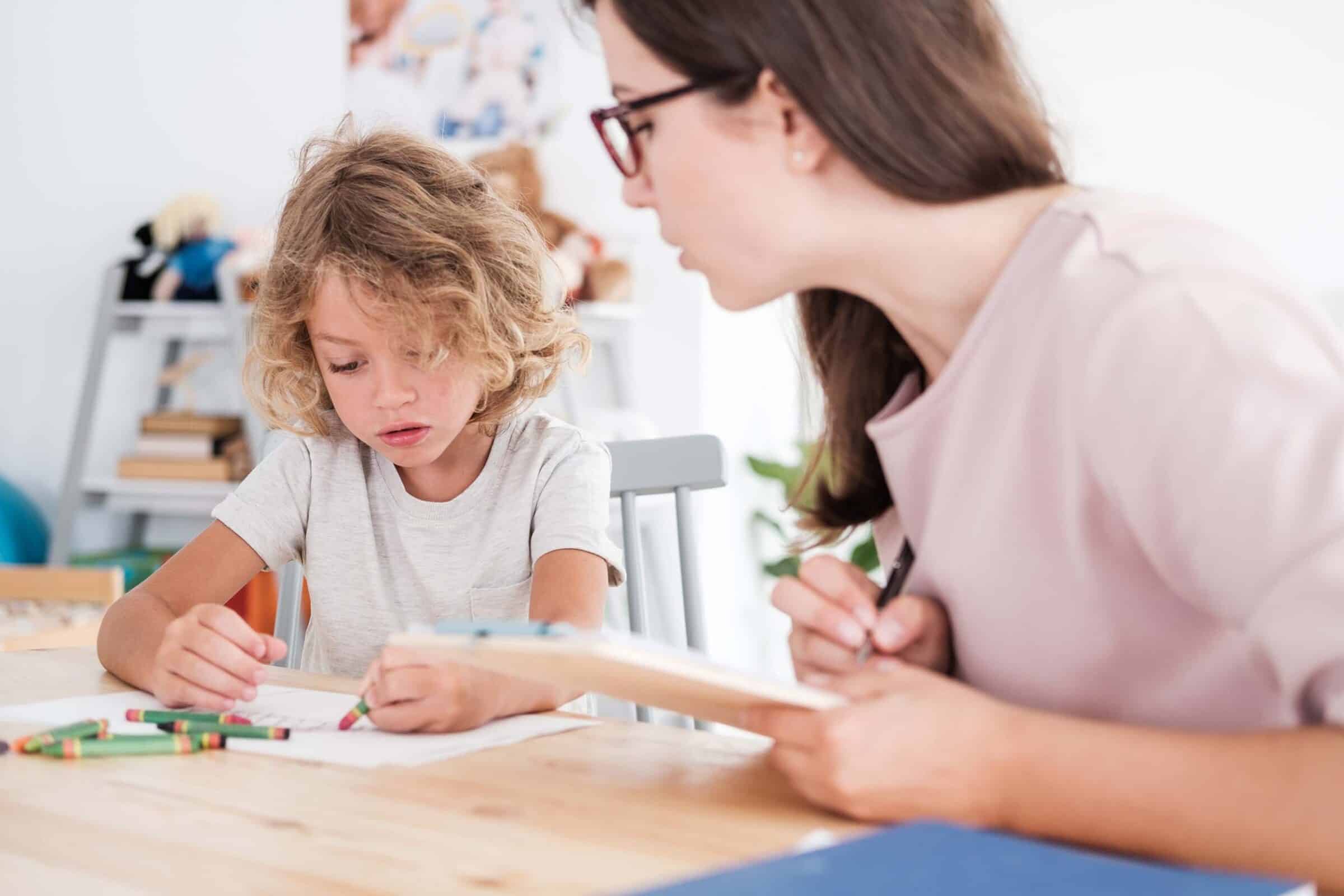 A psychotherapist watching a child draw pictures with crayons during an evaluation in a school.