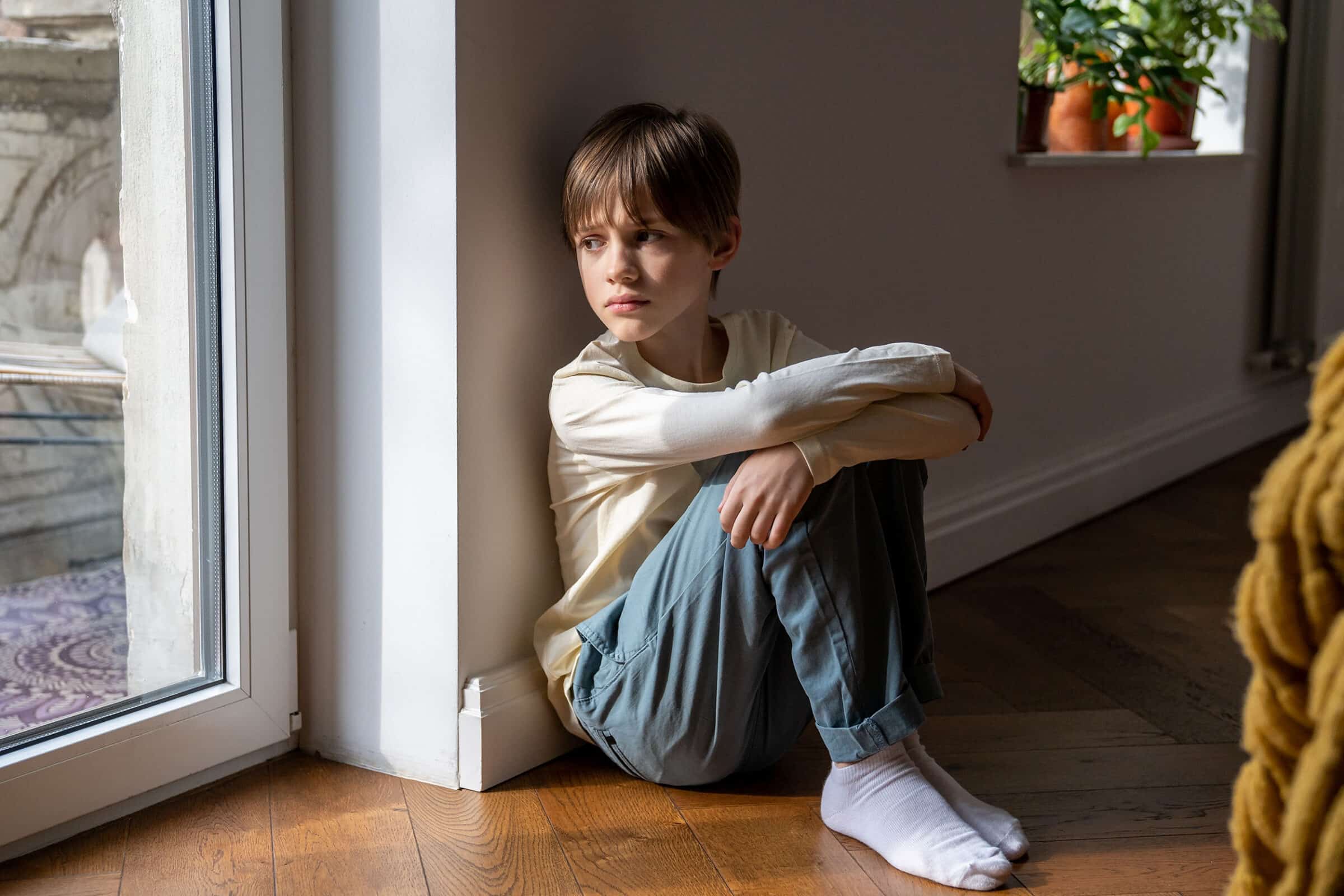 Kid psychological trauma. Lonely child boy sadly look to window hug knees sits on floor at home alone.