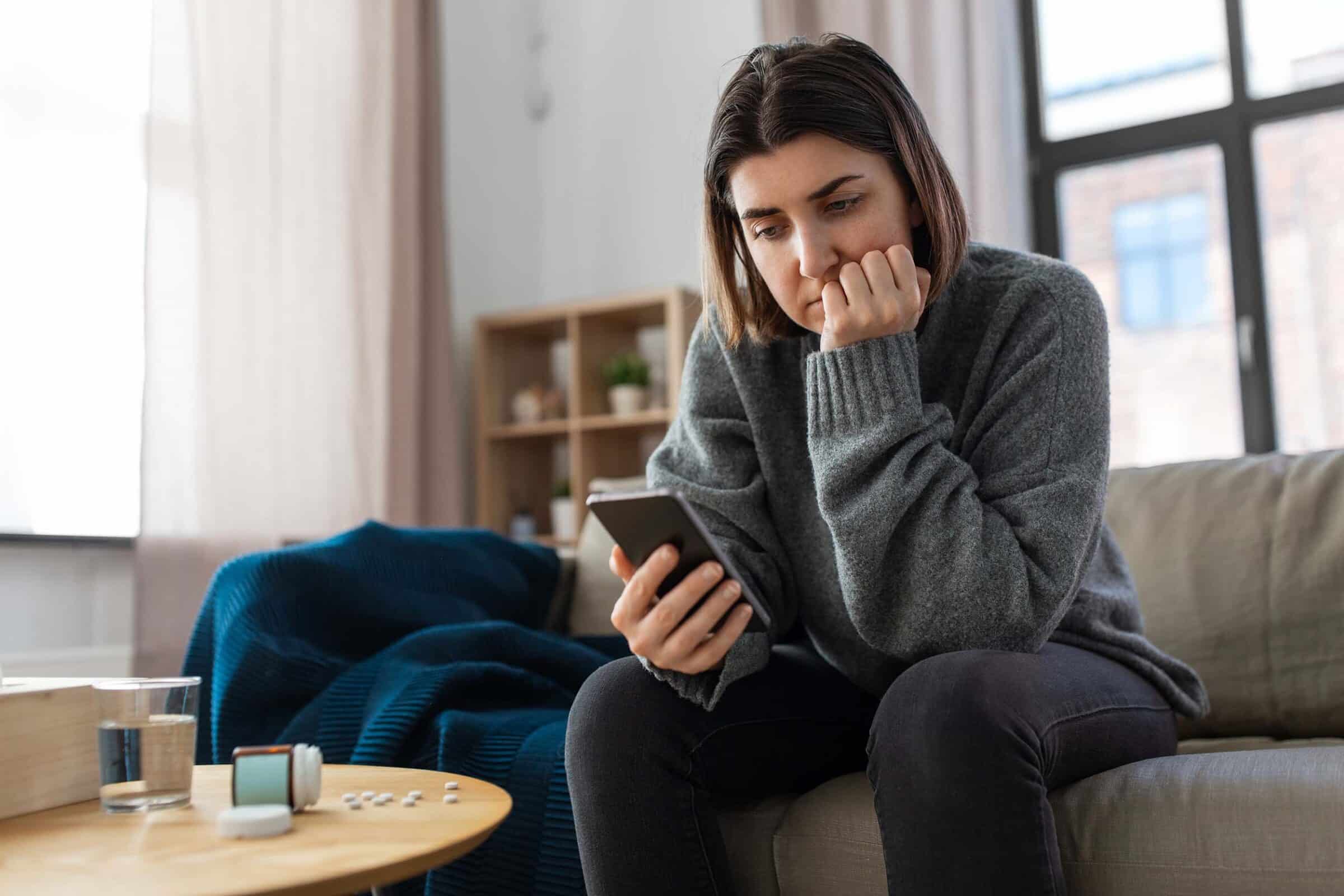 apathetic woman staring at her phone, medication is and water are on the table beside her
