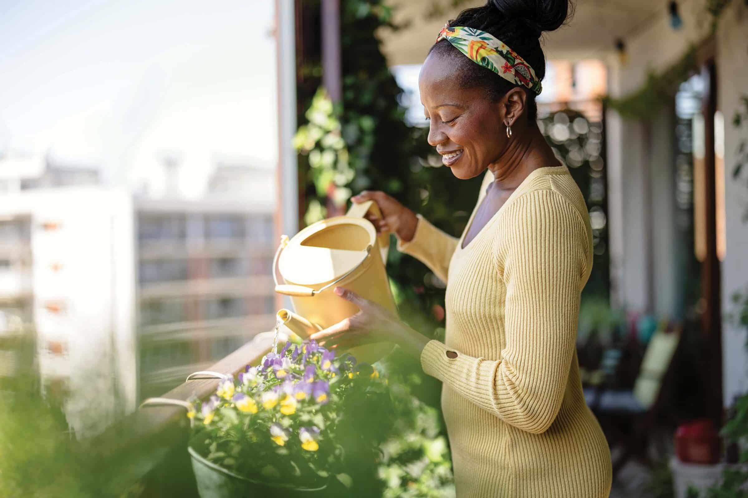 Content woman on her urban balcony, using a yellow watering can to tend to her outdoor container garden.