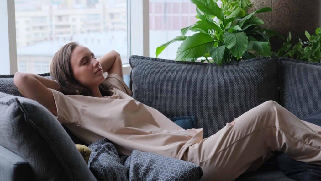 A calm young woman is resting on a couch with her eyes closed, her hands behind her head.
