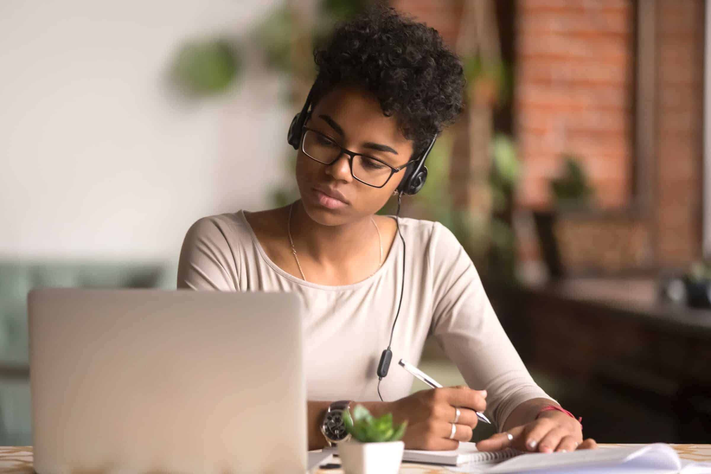 Focused multiracial woman wearing headphones write notes while studying on her laptop.